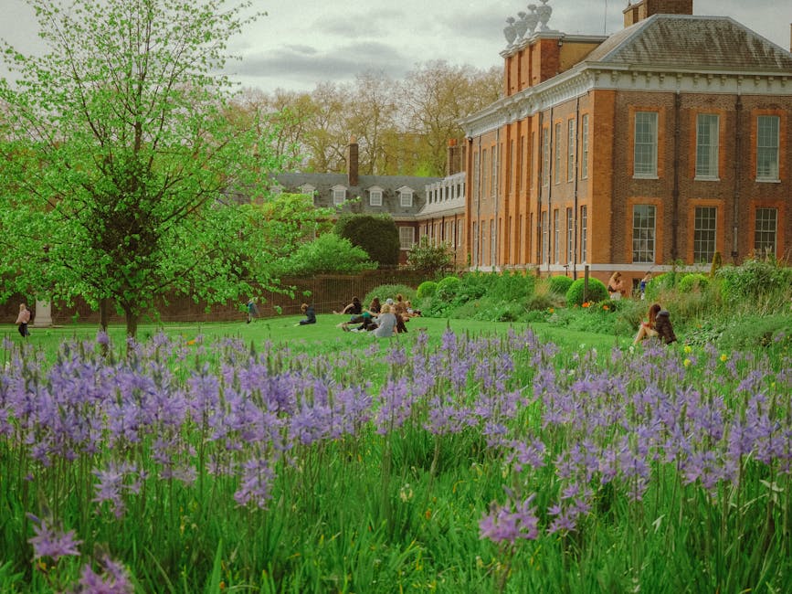 A lush green lawn with a variety of plants, including a large deciduous tree with bright green leaves, surrounds an historic brick building with white window frames and a hipped roof. In the foregroun